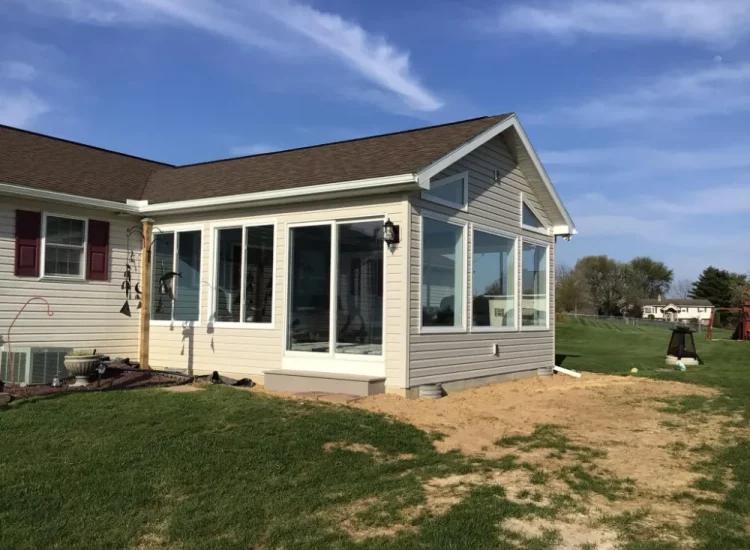 This image features the exterior of a contemporary sunroom extension attached to a house. The sunroom is characterized by its large windows, white siding, and a view of a vast, green landscape, making it a perfect relaxation spot.