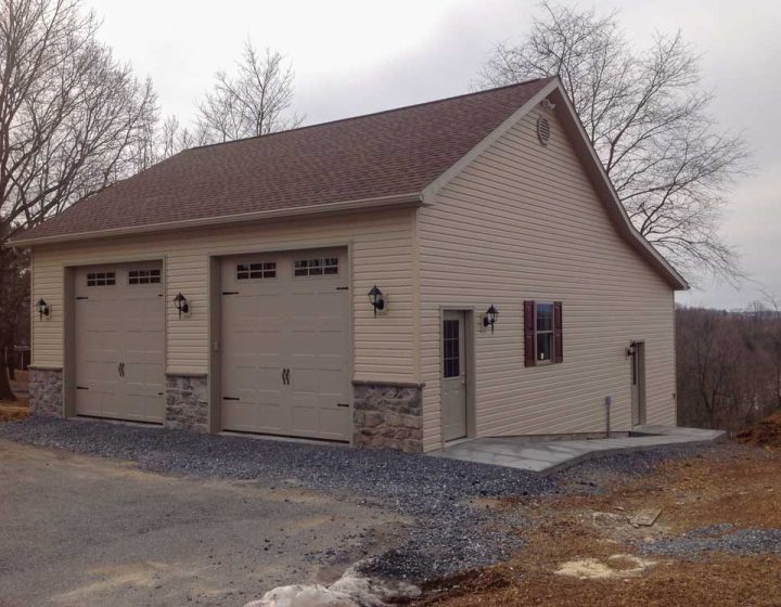 Modern garage building with beige siding and stone foundations, featuring two garage doors and practical lighting, set against a rural backdrop.