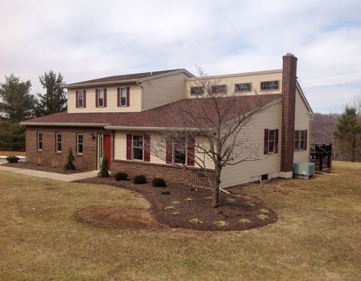 Exterior view of a two-story house featuring beige siding, red window shutters, and a brick facade, situated in a suburban setting with mature trees in the background.