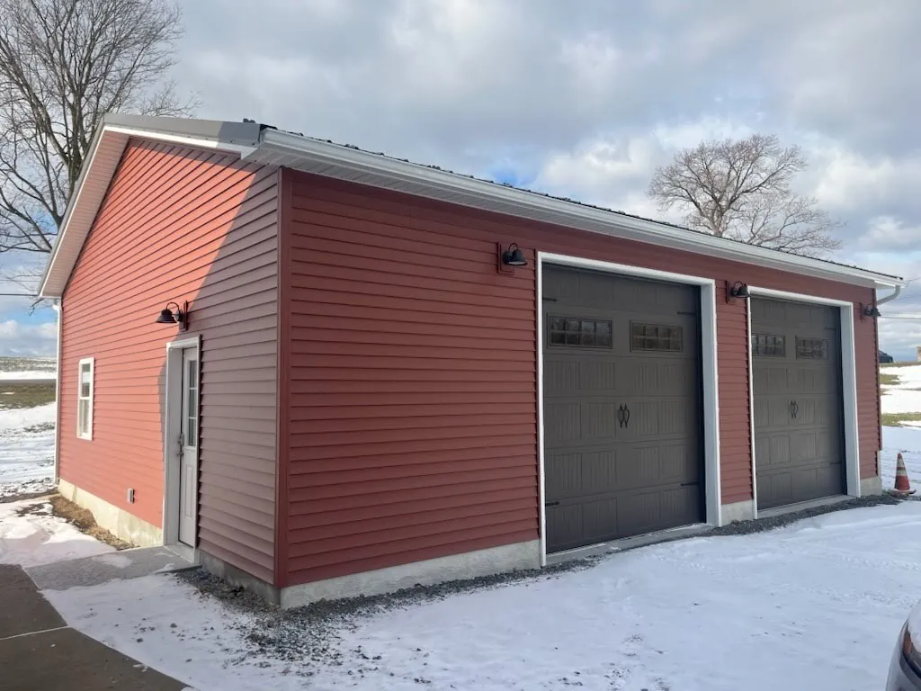 Two-car garage with red vinyl siding, brown carriage-style garage doors and barn light fixtures built by Leids Carpentry.